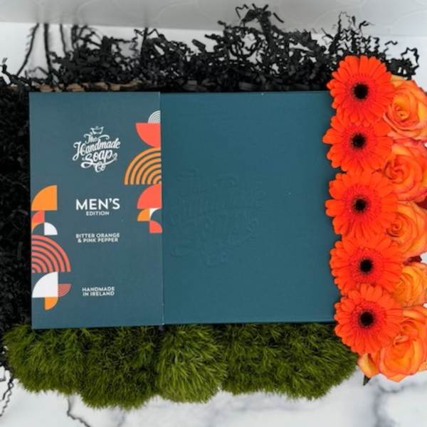 Men's soap gift box with bright orange gerbera daisies and roses, green moss and black filler in a tray