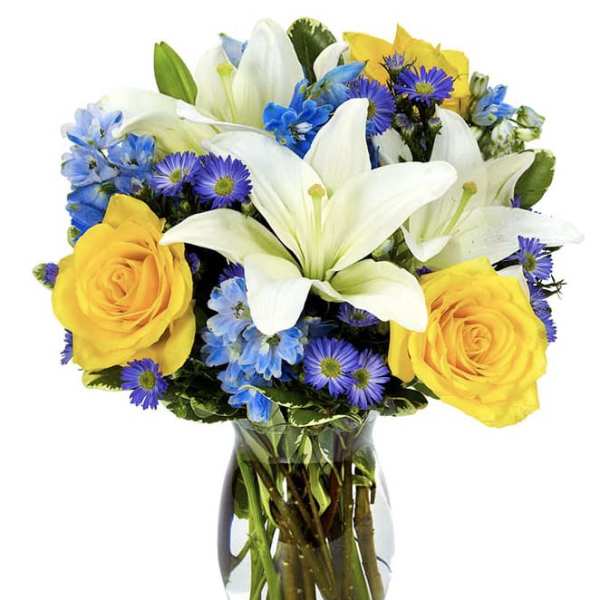 Bouquet of white lilies, yellow roses, and blue-purple flowers in a glass vase