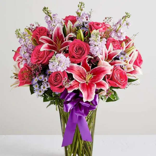 Pink roses and lilies arranged in a clear glass vase with a purple ribbon.