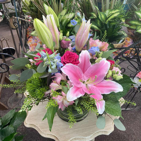 Mixed bouquet of pink lilies, roses, and accent blooms in a clear glass vase on a small wooden table