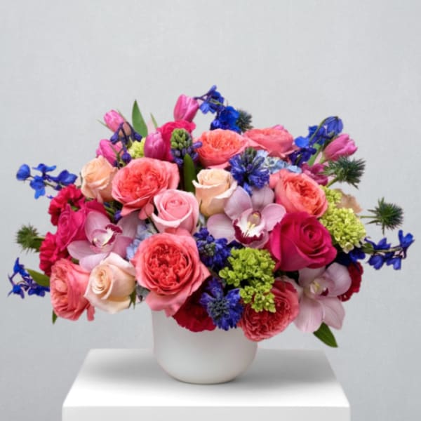Low arrangement of pink roses, blue delphinium, and mixed flowers in a white round vase