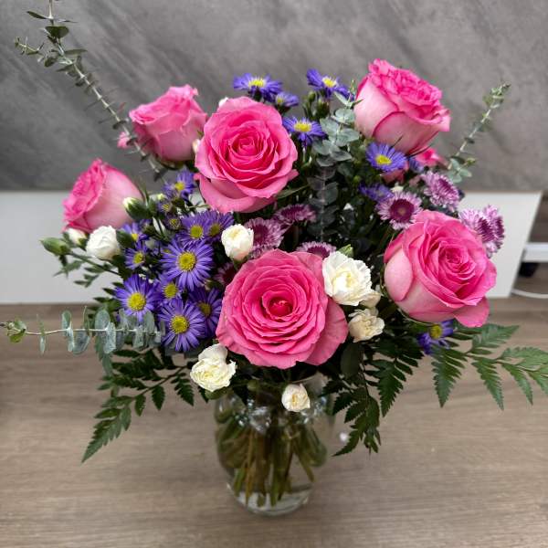 Arrangement of bright pink roses, purple asters, and white spray roses in a clear glass vase