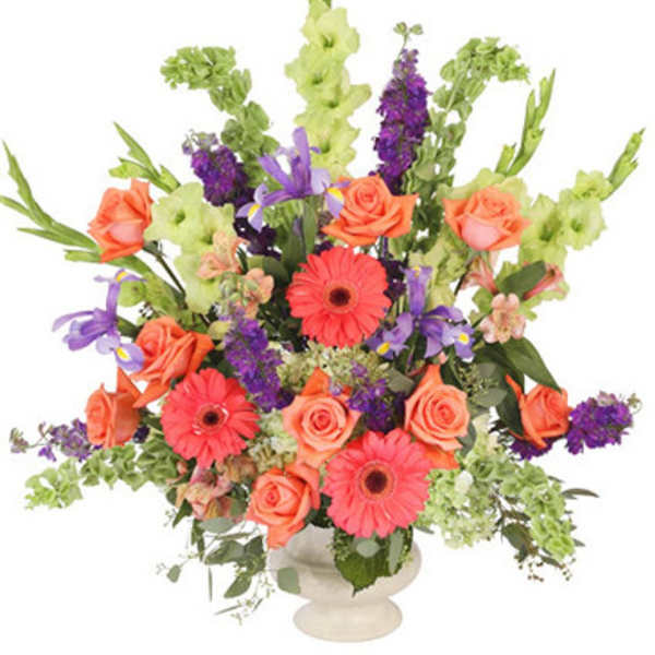 Tall arrangement of orange roses, gerbera daisies, purple blooms and green Bells of Ireland in a white pedestal vase