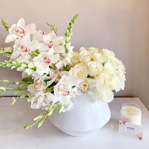 White roses and orchids arranged in a white vase beside a candle.