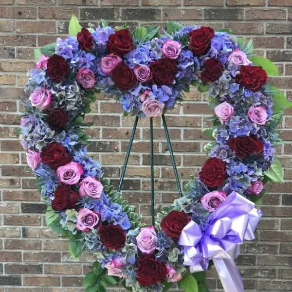 Heart-shaped standing wreath of red and pink roses with purple hydrangeas and a lavender bow on an easel.