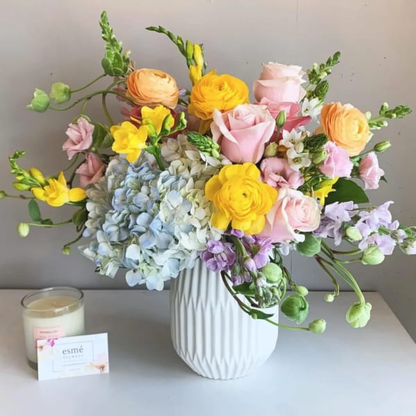 Mixed pastel bouquet in a white vase with a candle and card beside it
