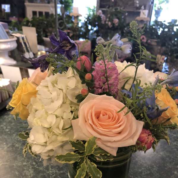 Low glass vase of peach roses, white hydrangeas, and mixed pastel flowers on a green countertop.