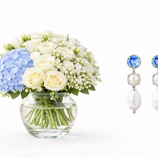 White roses and blue hydrangea in a glass vase beside blue-and-pearl earrings