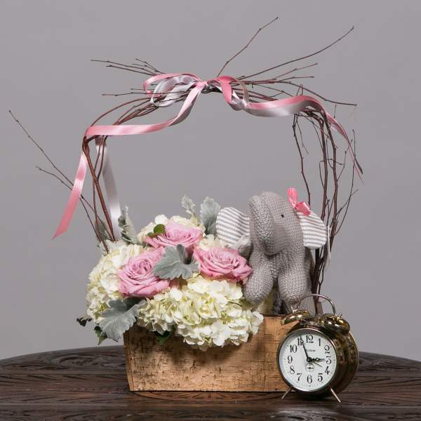 Pink roses and white hydrangeas in a wooden basket with a knitted elephant and clock