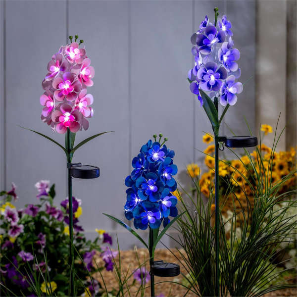 Set of three solar flower garden stakes with glowing pink, blue, and purple blooms in a garden bed.