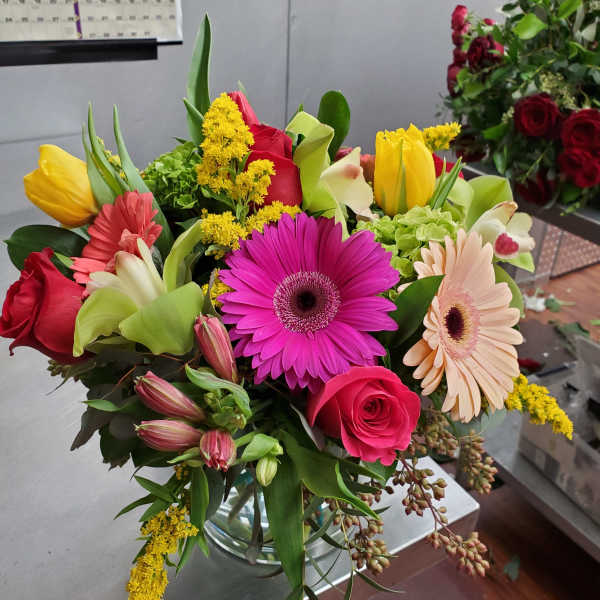 Mixed bouquet of pink gerbera daisies, roses, and yellow tulips in a clear glass vase on a worktable.