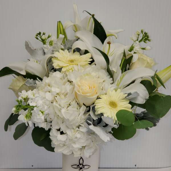 Tall white and cream flower arrangement with lilies, roses, hydrangeas and gerberas in a white vase with a silver cross.