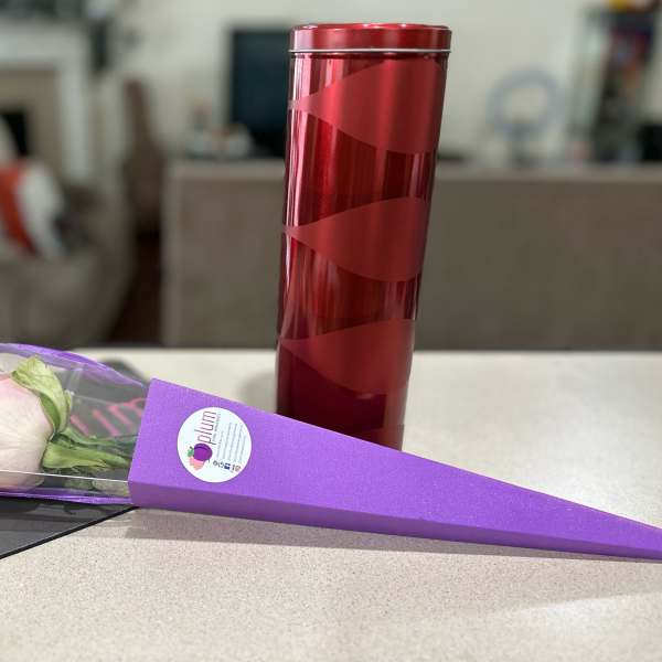 Single pale pink rose in a purple cone wrap beside a red cylindrical gift tin