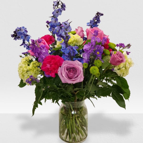 Colorful mixed bouquet of roses, carnations, delphinium, and other blooms in a clear glass vase.