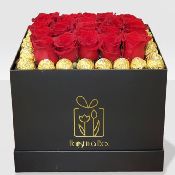 Square black box filled with red roses framed by gold-wrapped chocolates.