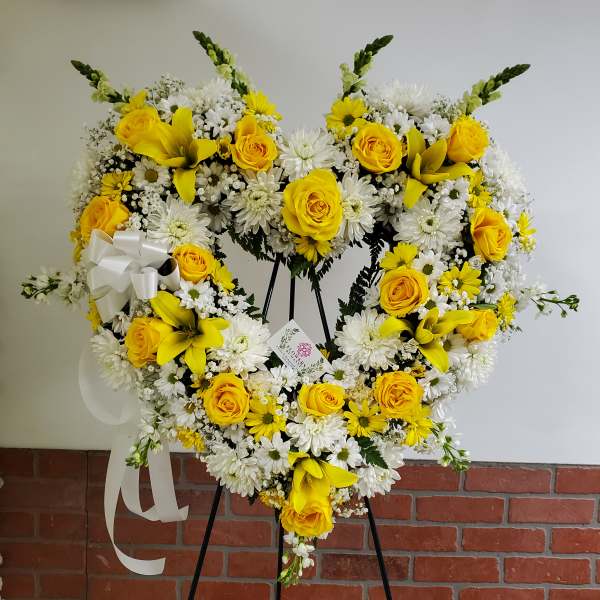 Heart-shaped standing wreath of yellow roses, white daisies and lilies with a white bow on an easel.