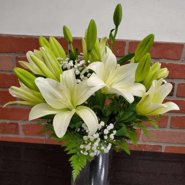 Tall arrangement of white lilies with buds and small white blooms in a glass cylinder vase