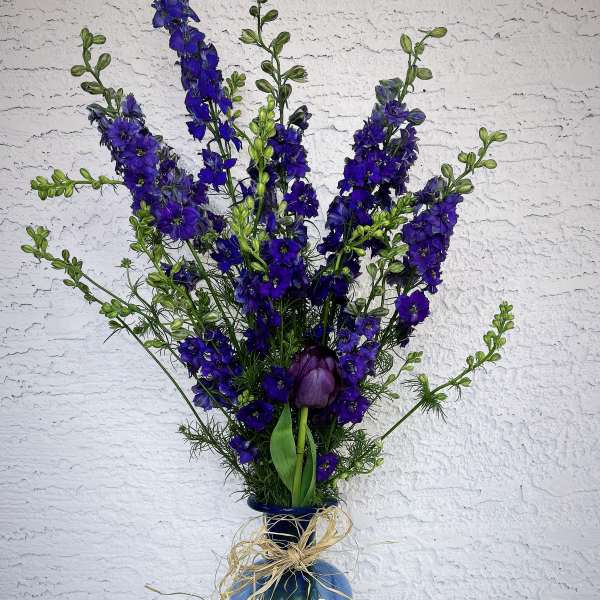 Tall blue floral arrangement in a glass vase with raffia bow