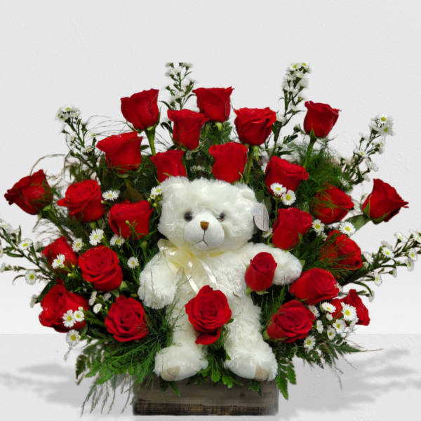 White teddy bear surrounded by red roses and small white flowers in a wooden box