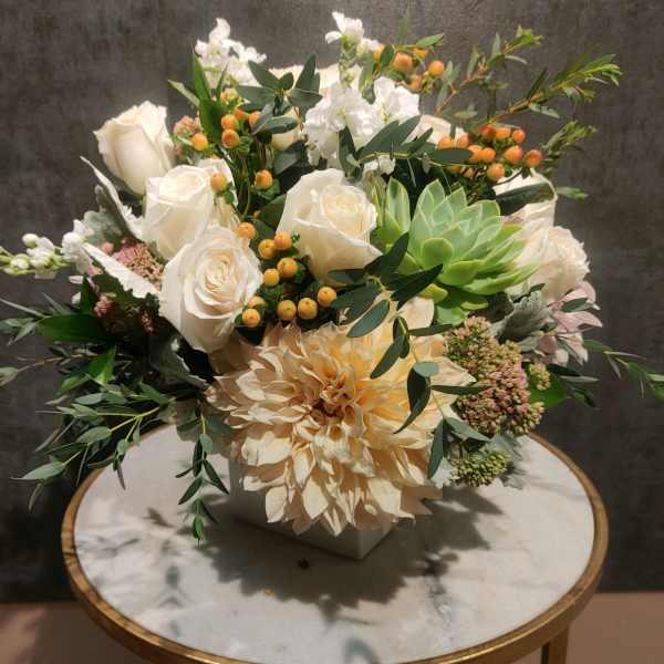White roses and a peach dahlia arranged in a square vase with greenery.
