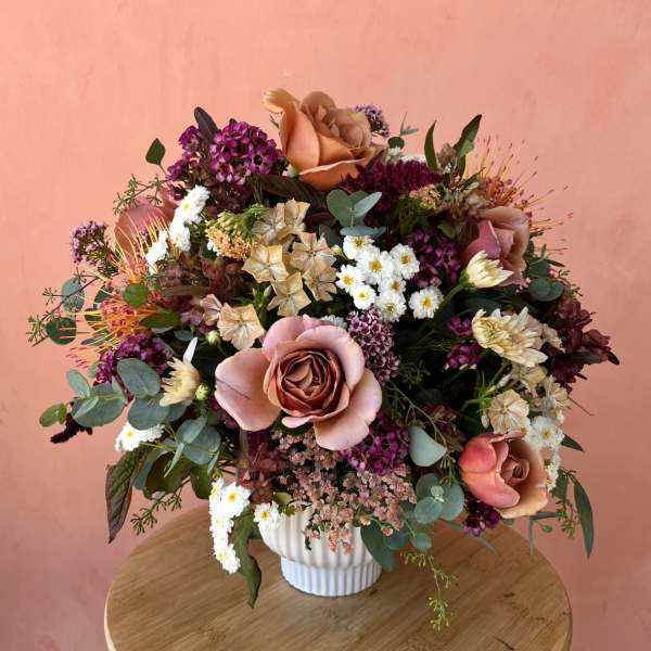 Mixed bouquet of blush roses, purple blooms, and white daisies in a white vase