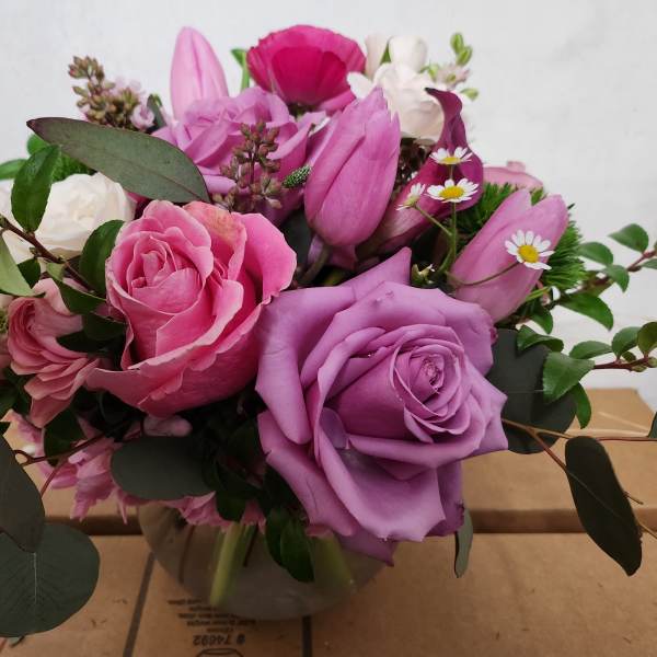 Low round arrangement of pink and lavender roses, tulips, and small white daisies in a clear glass bowl vase