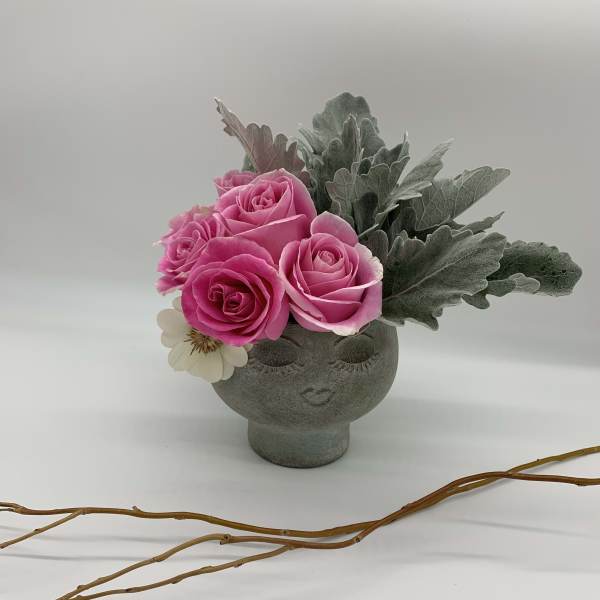 Pink roses arranged in a gray face-shaped vase with silvery foliage