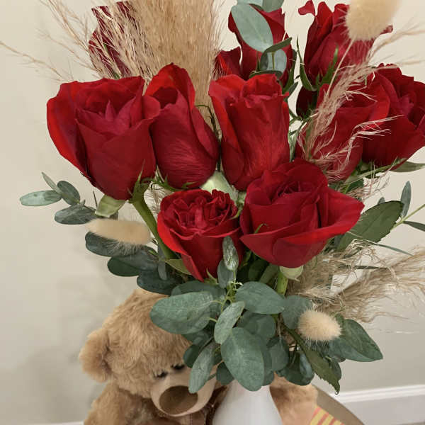 Red roses in a white vase with a teddy bear beside them