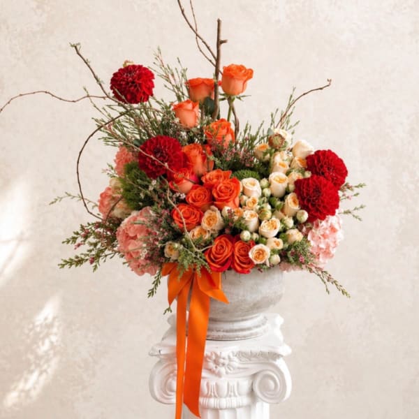 Orange and red rose arrangement in a white urn with an orange ribbon