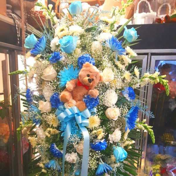 Tall floral standing spray with a teddy bear and blue ribbon