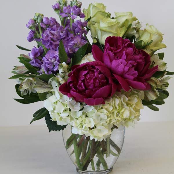 Mixed bouquet of white hydrangeas, purple stock, green roses, and magenta peonies in a glass vase