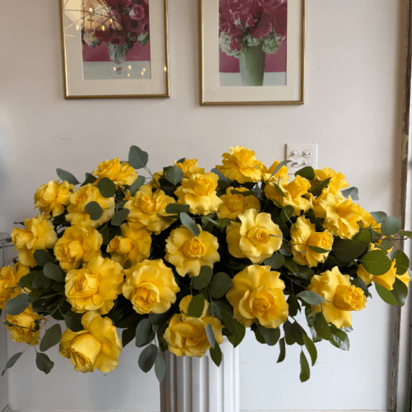 Large arrangement of yellow roses with round foliage in a white pedestal vase against a gallery wall