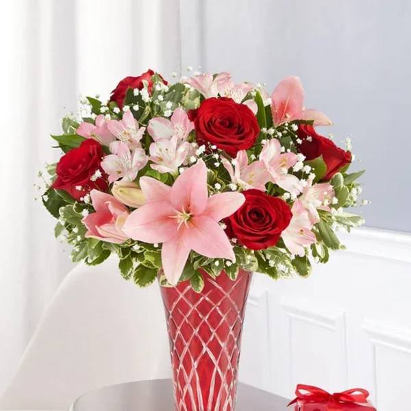 Red roses and pink lilies in a red glass vase with a small gift box