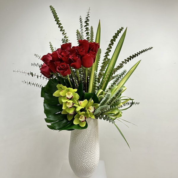 Tall white vase with red roses and green orchids arranged with tall linear foliage.