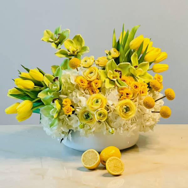 Yellow floral arrangement in a white bowl with lemons in front