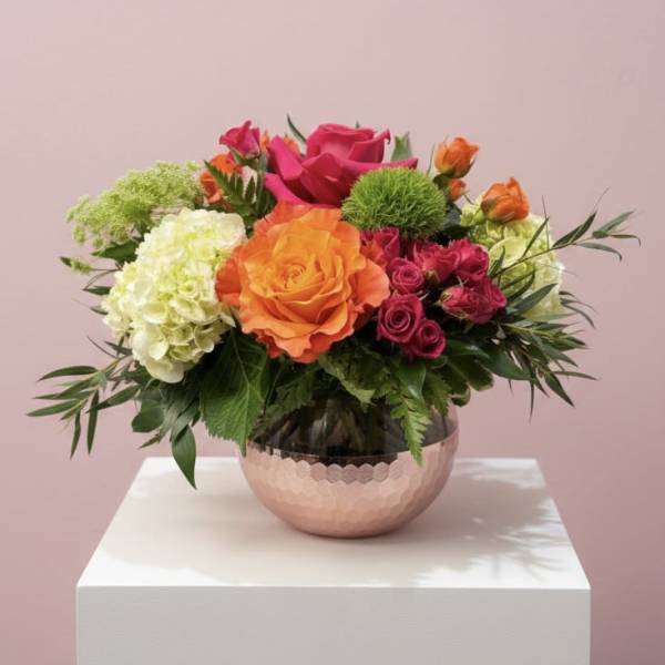 Bright orange, pink, and white flowers in a low hammered copper bowl vase on a white pedestal