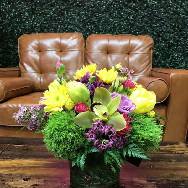 Low glass vase of yellow mums, purple blooms, green dianthus, and an orchid on a wooden table