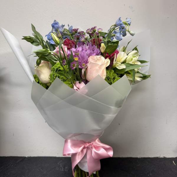 Mixed bouquet of pink, purple, blue, and white flowers wrapped in white paper with a pink ribbon