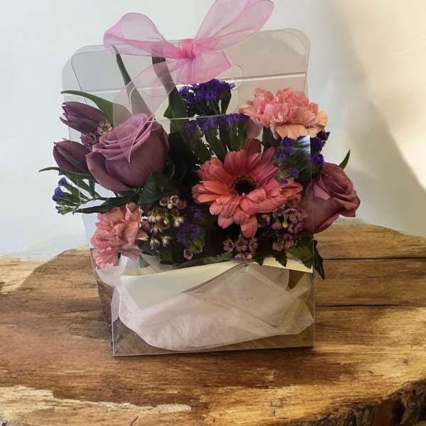 Pink and purple flower arrangement in a clear gift box with a ribbon bow
