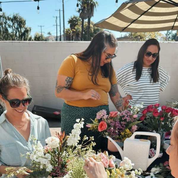 People arranging flowers at an outdoor table under an umbrella.