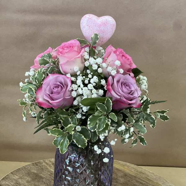 Pink roses with baby's breath in a purple glass vase, topped with a heart pick