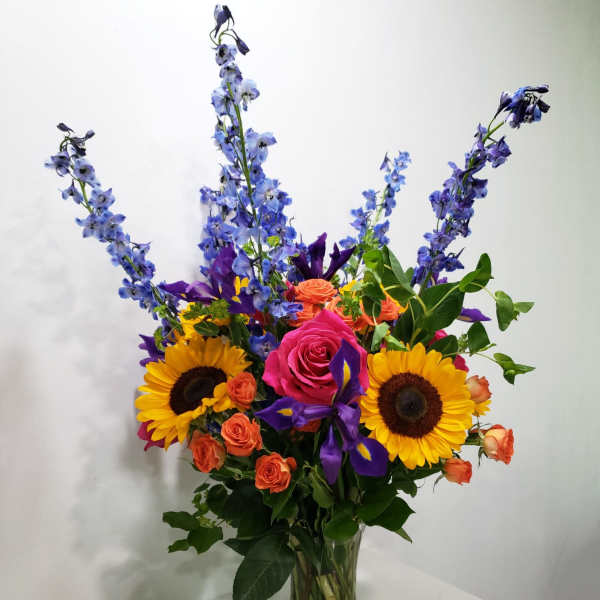 Mixed bouquet in a clear glass vase with sunflowers, roses, and blue delphinium