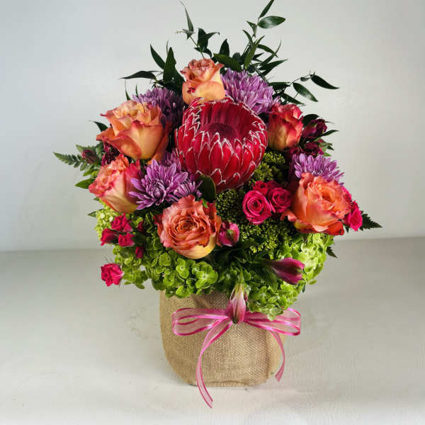 Mixed bouquet with roses, purple mums, and a red protea in a burlap-wrapped container