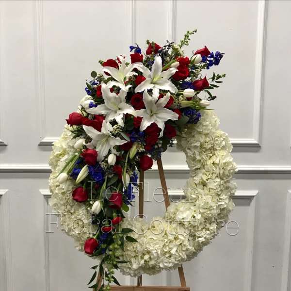 Large floral wreath on an easel with red roses, white lilies, and blue accents