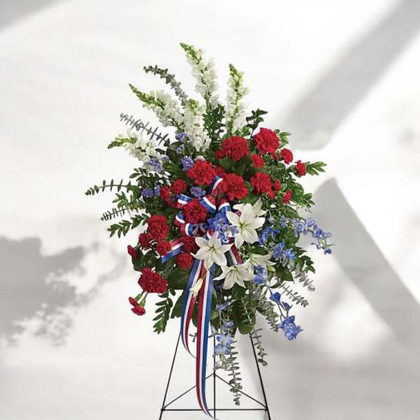 Standing floral spray with red carnations, white lilies, and blue accents on a metal easel