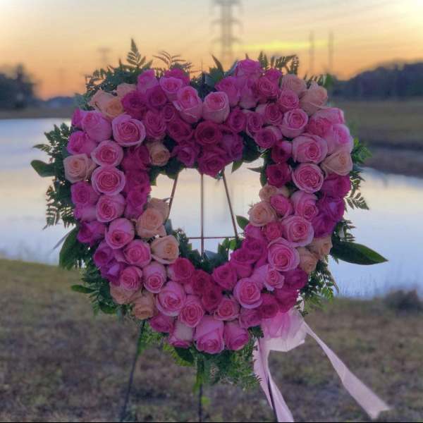 Heart-shaped standing wreath of pink and peach roses on an easel with trailing ribbons outdoors at sunset