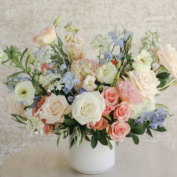 Pastel roses, lisianthus, and blue delphinium in a white ceramic vase on a light table
