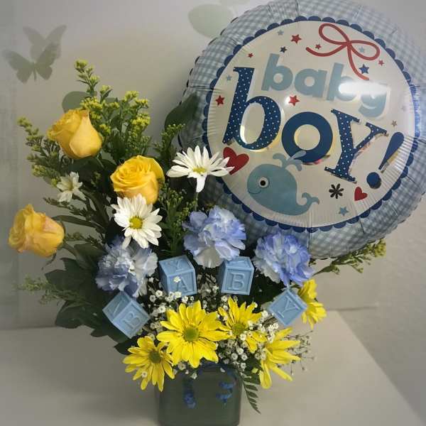 Baby boy-themed bouquet with yellow roses, blue carnations, daisies, and a baby boy balloon in a green cube vase