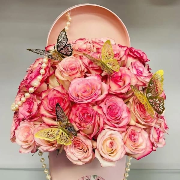 Pink roses arranged in a round pink hat box with butterfly accents and pearl strands.