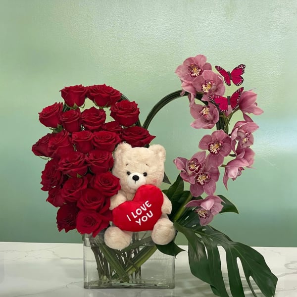 Red roses and pink orchids in a glass vase with a teddy bear holding a heart